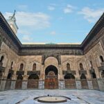 Interior courtyard of Madrasa Bou Inania in Fez, Morocco, featuring ornate Islamic architecture, intricate tilework, and a central marble fountain.