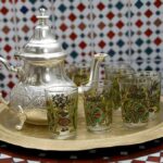 Moroccan Tea Ceremony : Discover the Fascinating Traditions Behind This Beloved Ritual 6 A shiny silver Moroccan teapot and several ornate glasses sit on a round brass tray with intricate etchings, all placed on a table with a blue and white geometric tile pattern, against a backdrop of colorful patterned tiles.